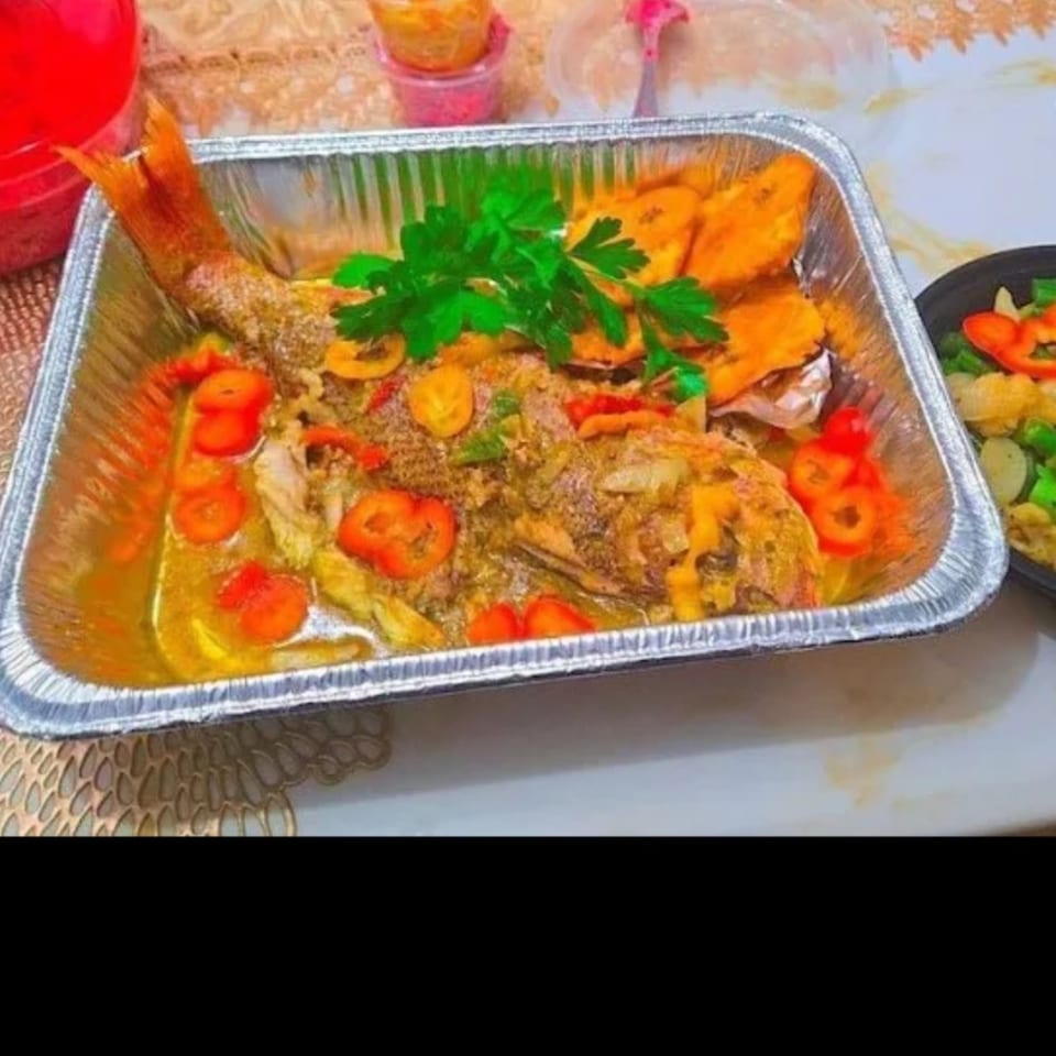 Whole baked fish garnished with parsley and sliced peppers, served with roasted vegetables in a foil tray.