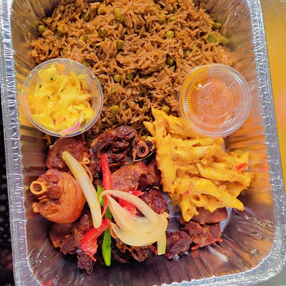 Aluminum takeout tray with spiced rice and peas, grilled meat, yellow pasta, pickled vegetables, and two dipping sauces.