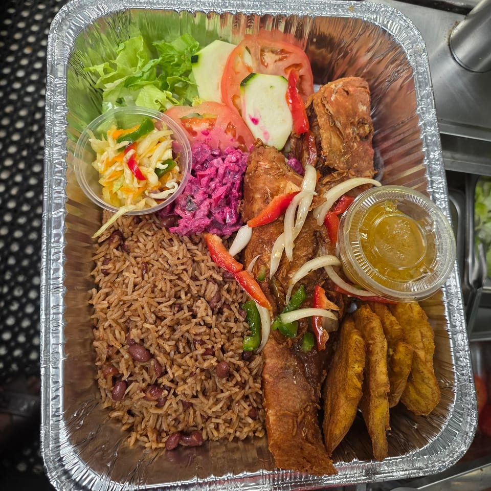 Aluminum takeout tray with rice and beans, fried plantains, grilled meat, salad, pickled vegetables, and dipping sauces.