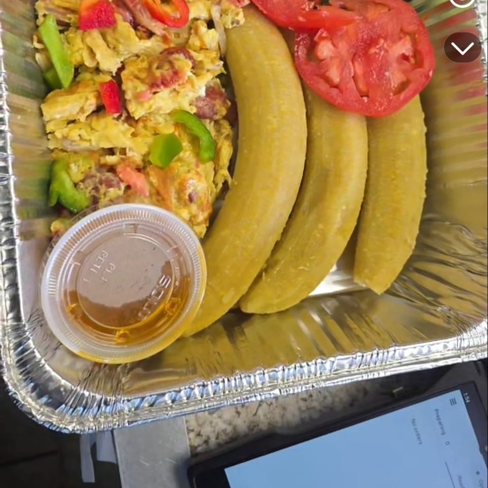 Aluminum takeout tray with scrambled eggs, fried plantains, fresh salad, sliced tomatoes, cucumbers, peppers, and dipping sauce.