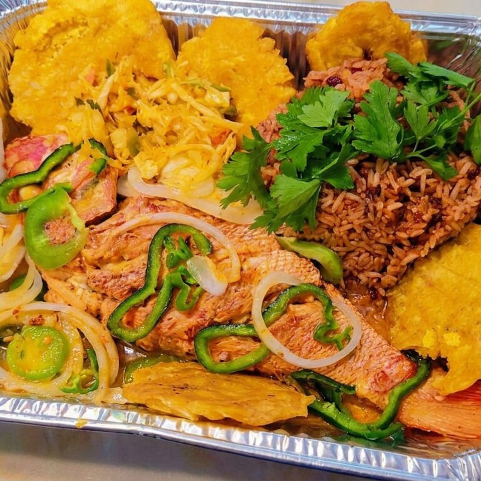 Aluminum tray of seasoned rice, grilled fish, fried plantains, vegetables, peppers, onions, and fresh parsley.