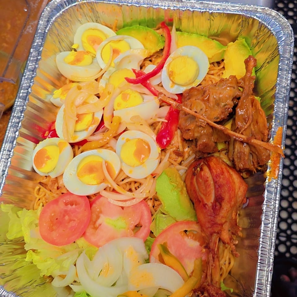 Aluminum takeout tray filled with noodles, sliced boiled eggs, lettuce, tomatoes, onions, peppers, and grilled meats.